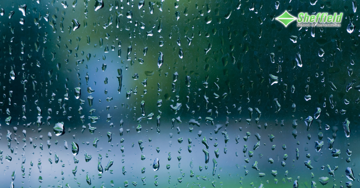 A piece of glass is covered in rain drops. Through the glass and water, blurred images of items that are green and blue can be seen. It appears the perspective is someone standing inside looking out their window. This relates to the article Does Rain Cancel Flights? 